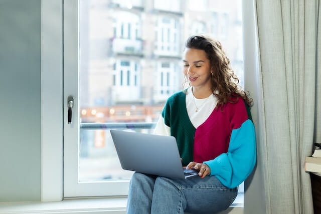 vrouwelijk persoon zit vrolijk met laptop bij het raam te werken aan blog